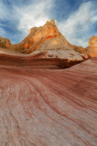White Pocket sandstone formation with red and white striations leading to sunlit cliffs under a dramatic sky.