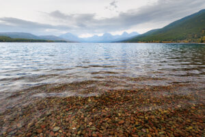 Lake McDonald shoreline with colorful stones under clear water, mountains in background, Glacier National Park landscape photography