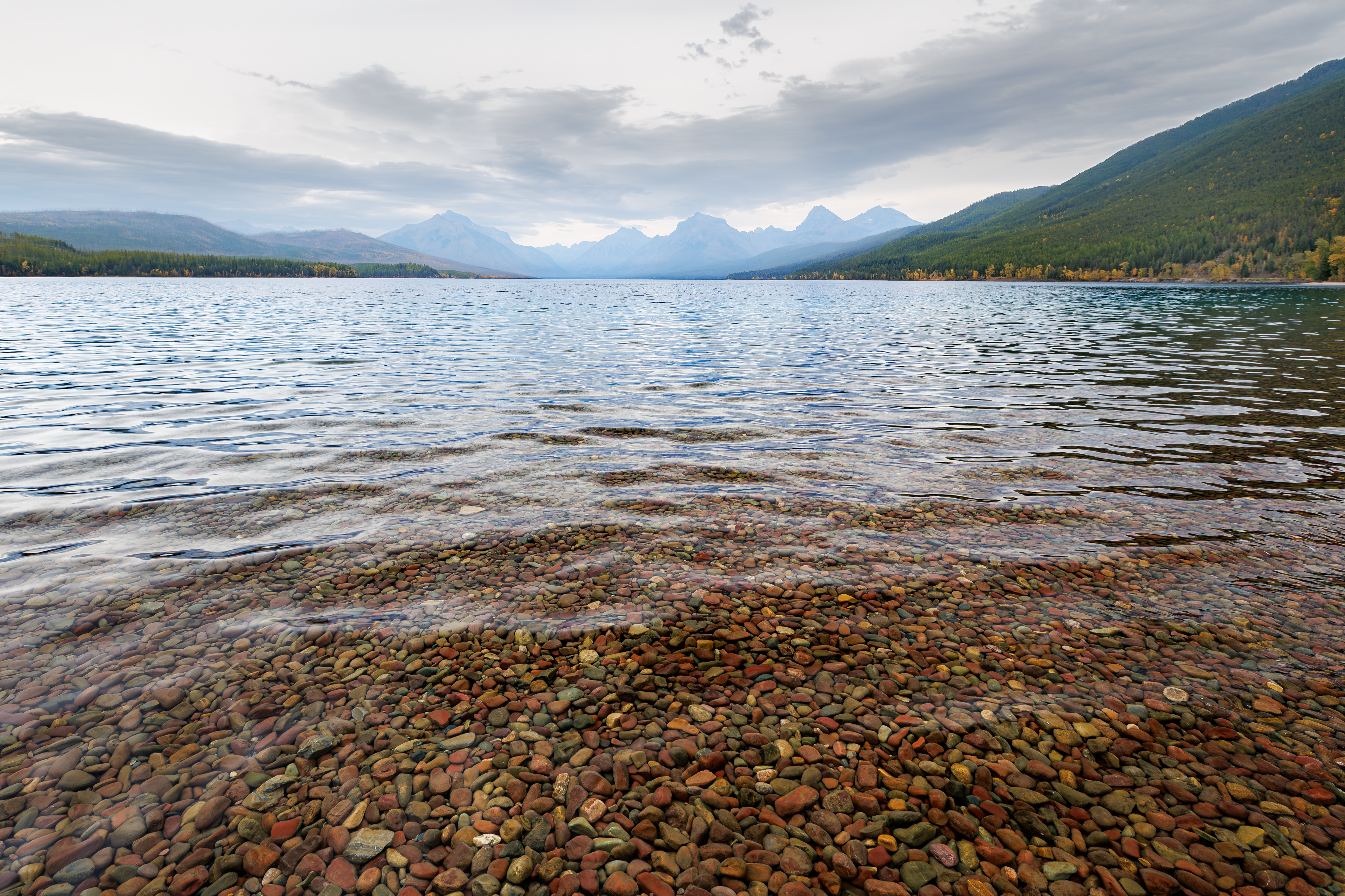 Lake McDonald shoreline with colorful stones under clear water, mountains in background, Glacier National Park landscape photography