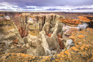 Panoramic view of Coal Mine Canyon in northern Arizona, featuring tall sandstone spires and colorful rock formations under a dramatic cloudy sky.